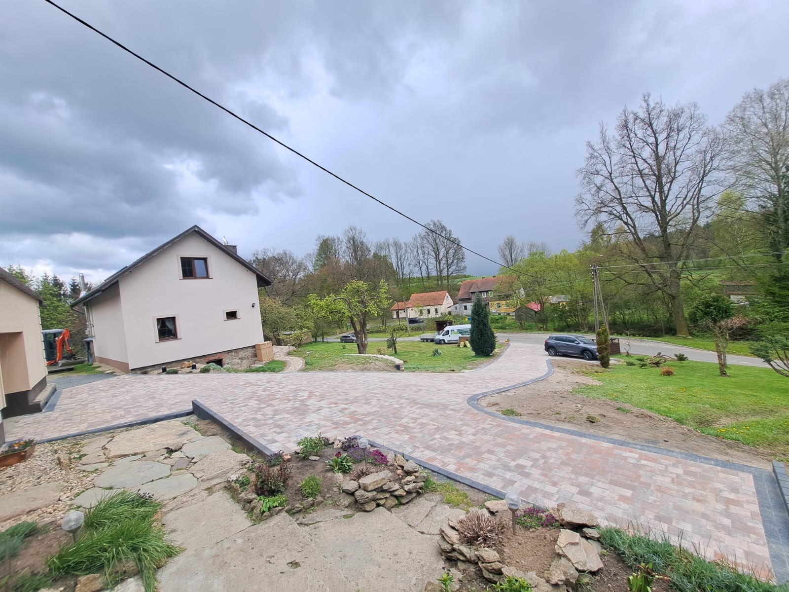 Nowo wyłożony, zakrzywiony podjazd z kostki brukowej w odcieniach czerwieni i szarości, prowadzący do domu z kremową elewacją, widok z góry z fragmentem kamiennych schodów i rabatki kwiatowej...