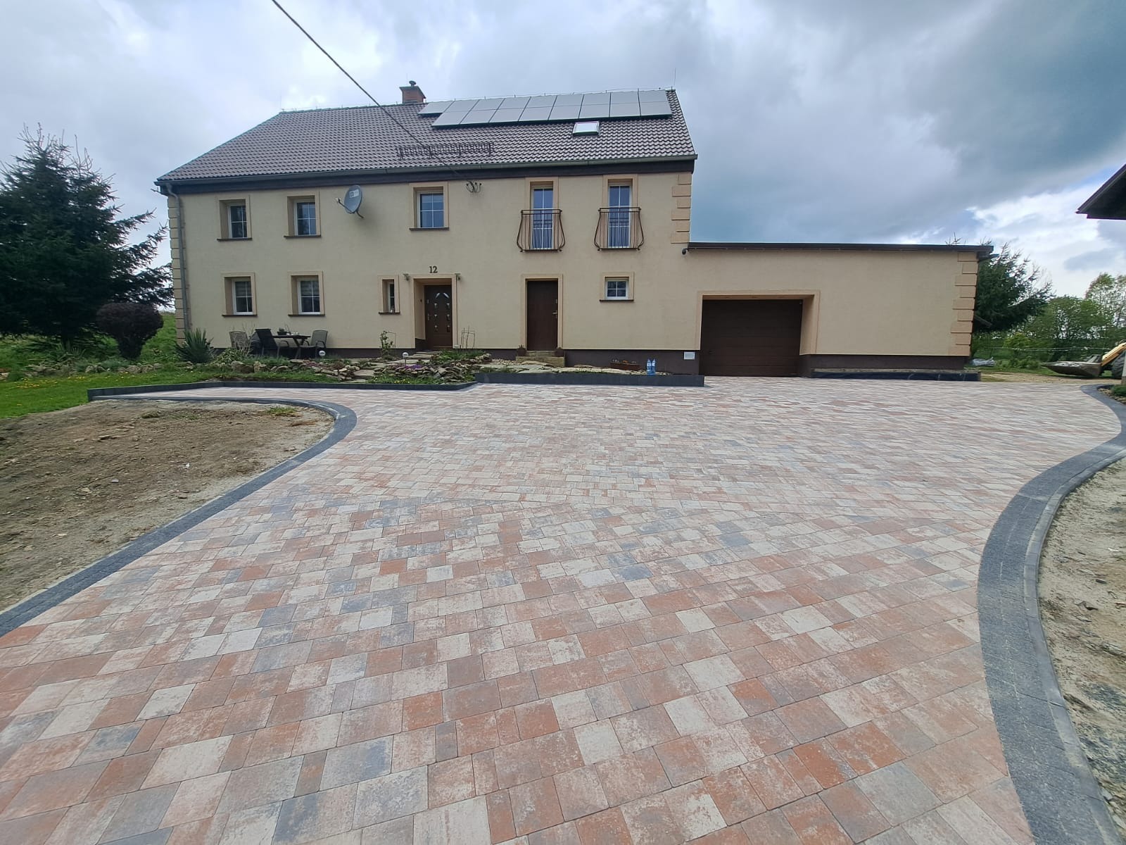 Nowo ułożona, wielokolorowa kostka brukowa prowadząca do beżowego domu z panelami słonecznymi na dachu, widoczne ślady po niedawnych pracach ziemnych po lewej stronie.