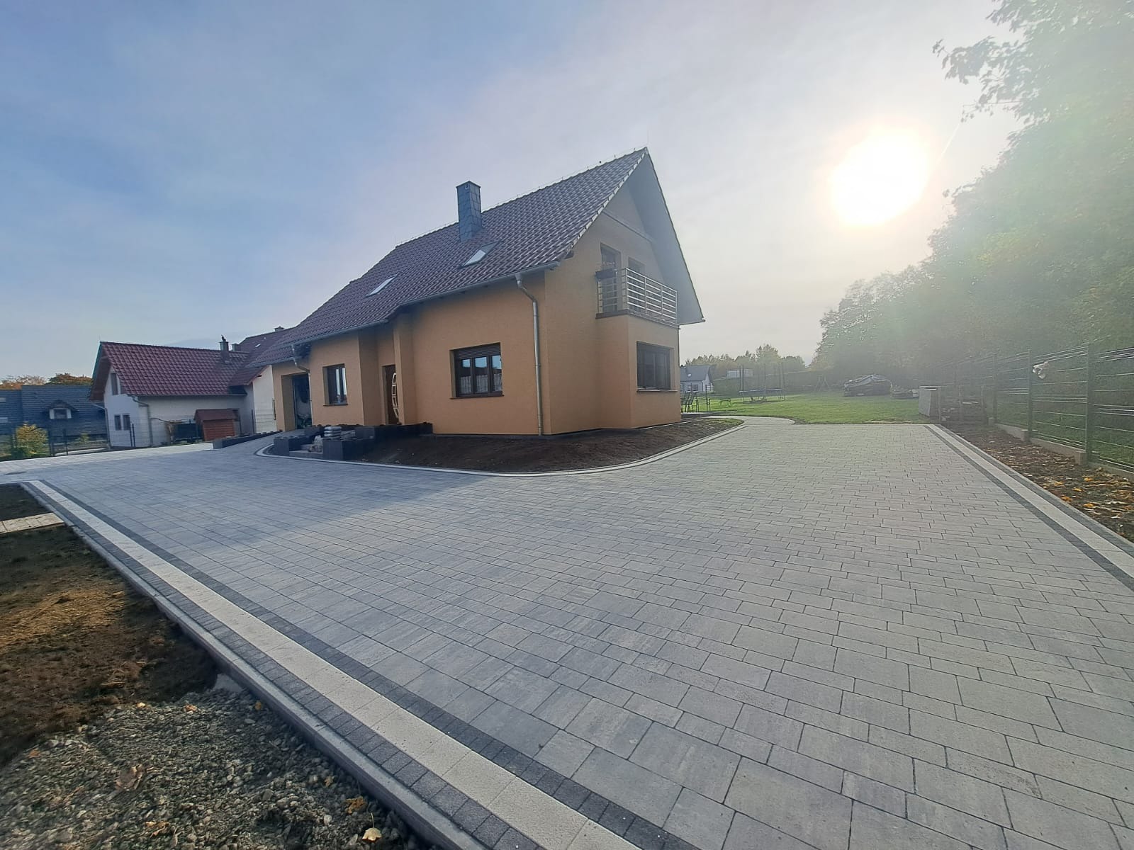 Nowo ułożony, rozległy podjazd z szarej kostki brukowej prowadzący do domu z beżową elewacją, widoczne obrzeża z ciemniejszej kostki, świeżo obsypana ziemia wokół budynku, w tle trawnik i drzewa.