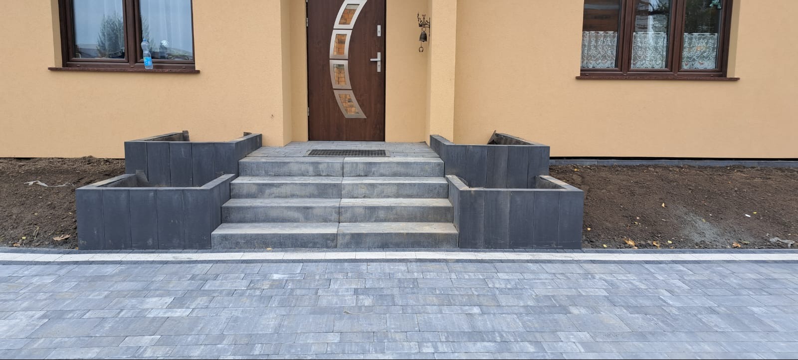 Schody zewnętrzne z szarej kostki brukowej prowadzące do brązowych drzwi wejściowych, z donicami po bokach i fragmentem ułożonej nawierzchni z kostki.