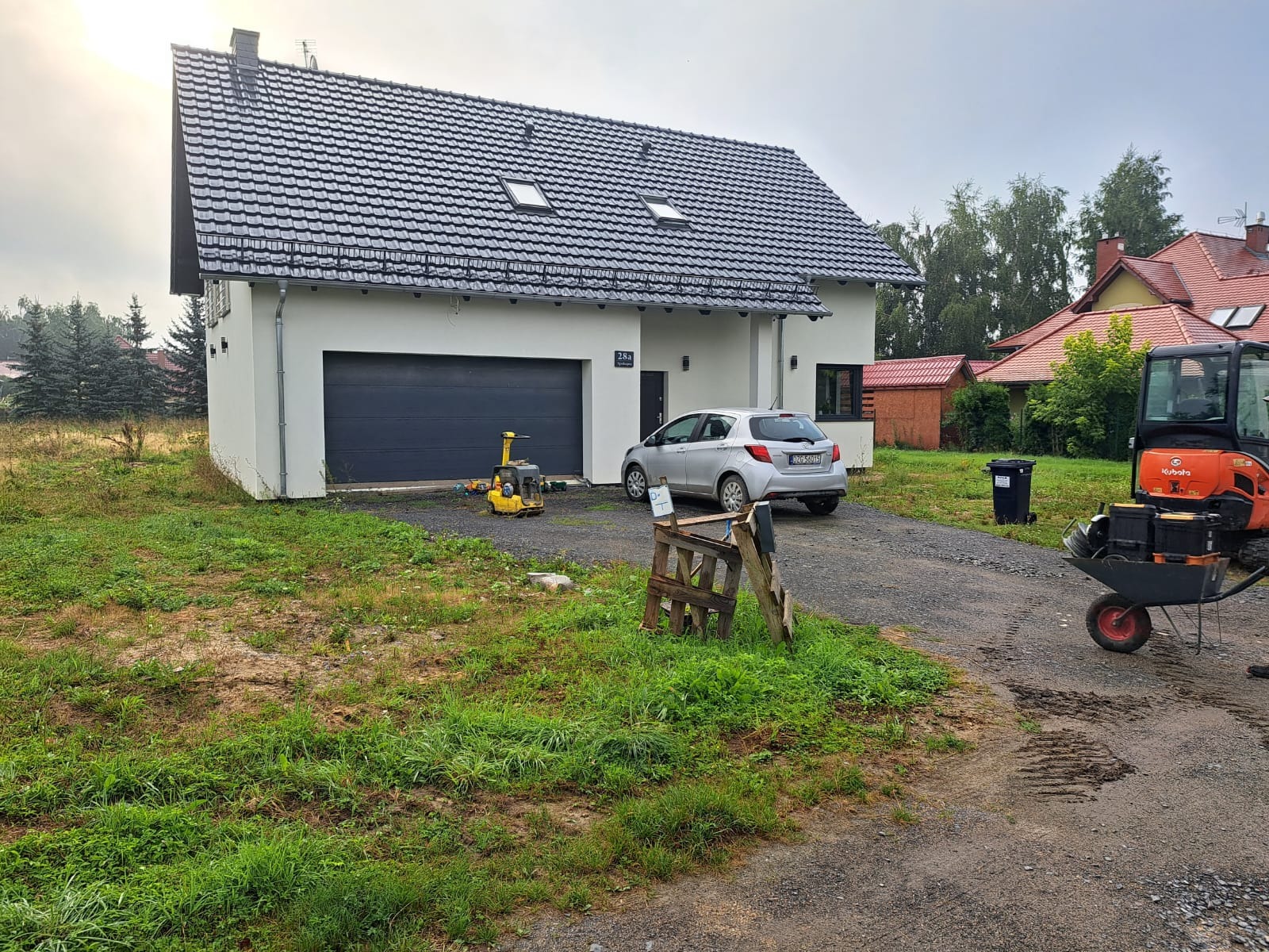 Przygotowanie terenu pod budowę: zagęszczarka wibracyjna i minikoparka Kubota na żwirowej drodze dojazdowej przed nowoczesnym domem z szarą bramą garażową i trawnikiem w trakcie niwelacji.