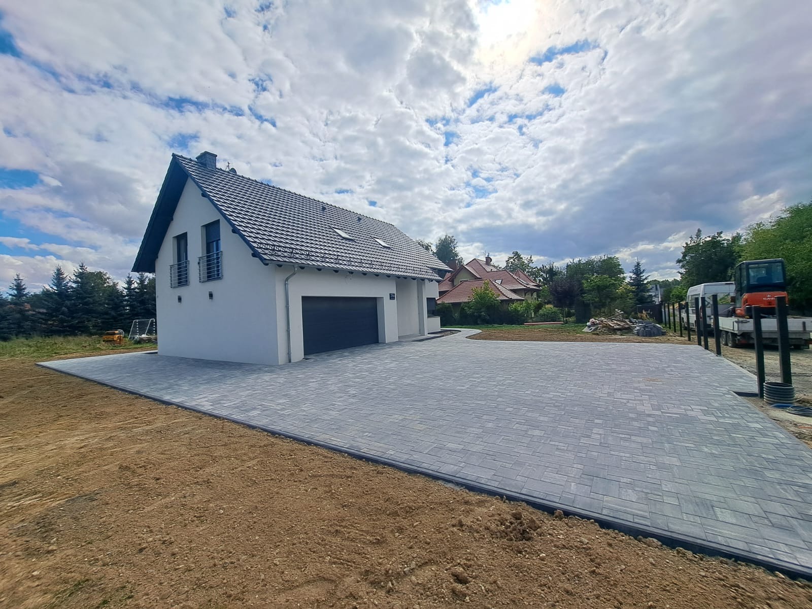 Nowoczesny podjazd z szarej kostki brukowej ułożonej we wzór jodełki, obrzeża z palisady, widok na biały dom z garażem i przygotowany teren pod trawnik.