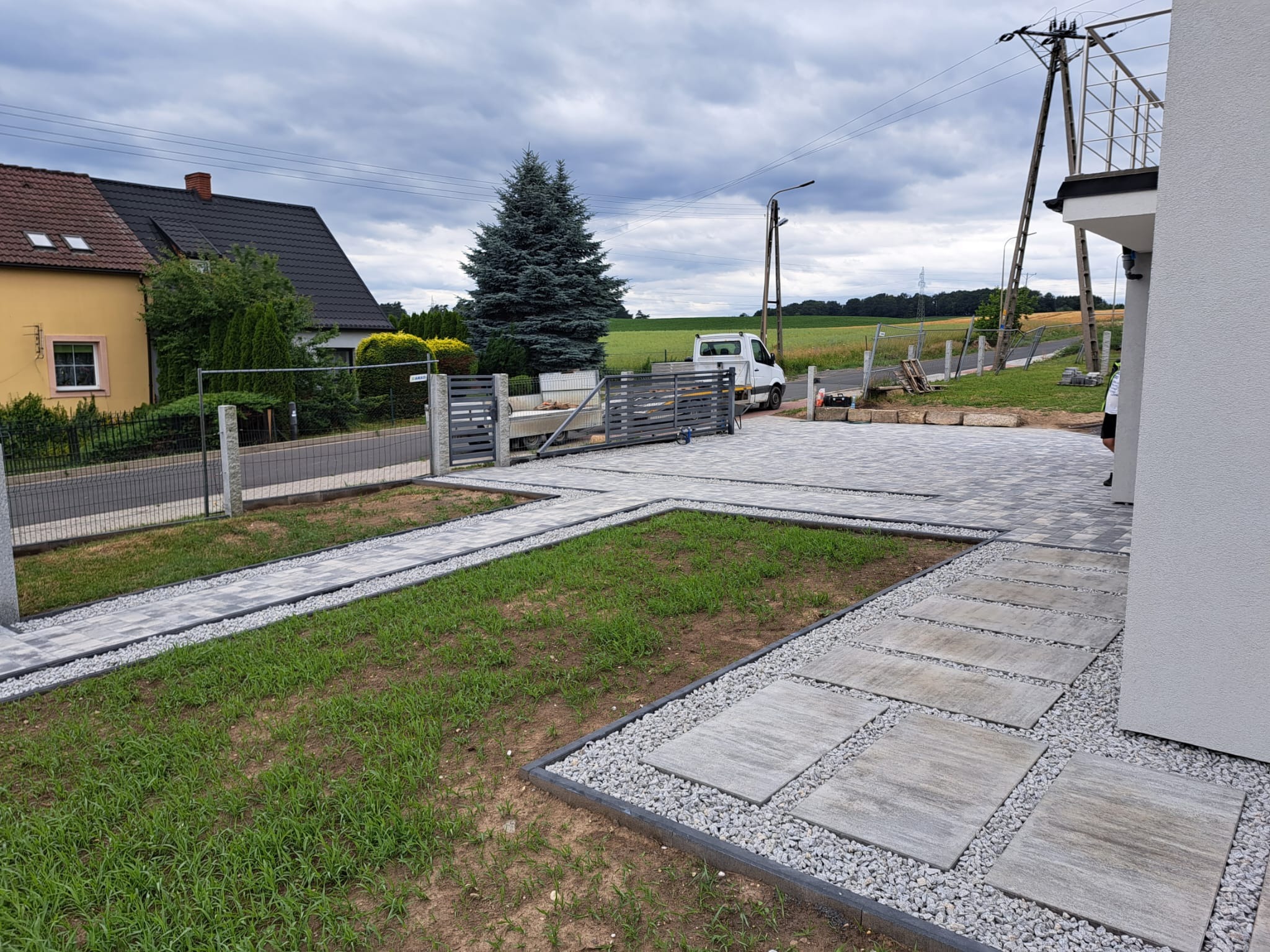 Ułożona kostka brukowa w geometryczny wzór na podjeździe, z elementami trawnika i ozdobnego żwiru, brama w tle, pochmurne niebo.