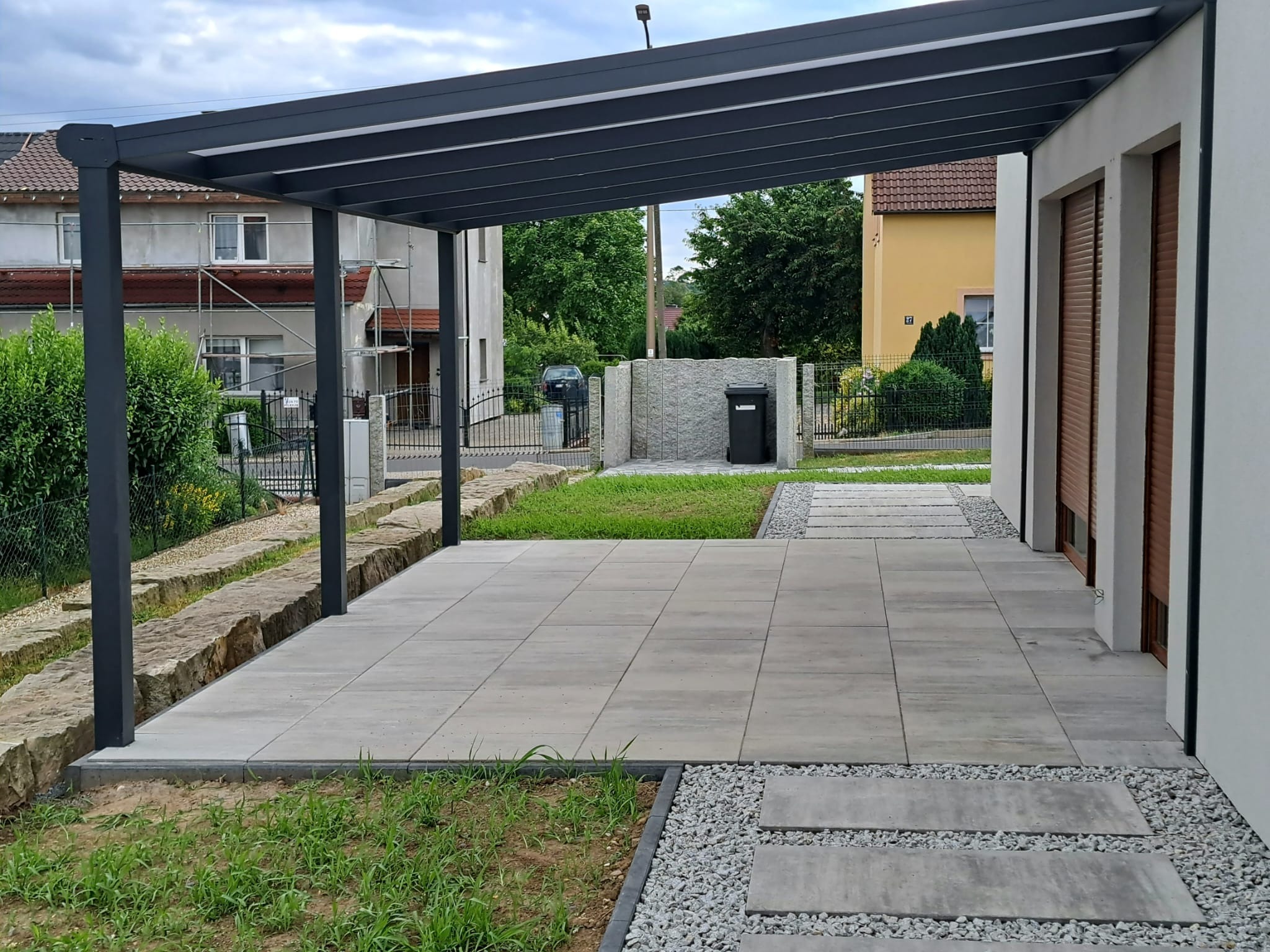 Nowoczesny taras z szarych płyt betonowych pod metalową pergolą, obrzeża z drobnego grysu, w tle domy jednorodzinne i trawnik.