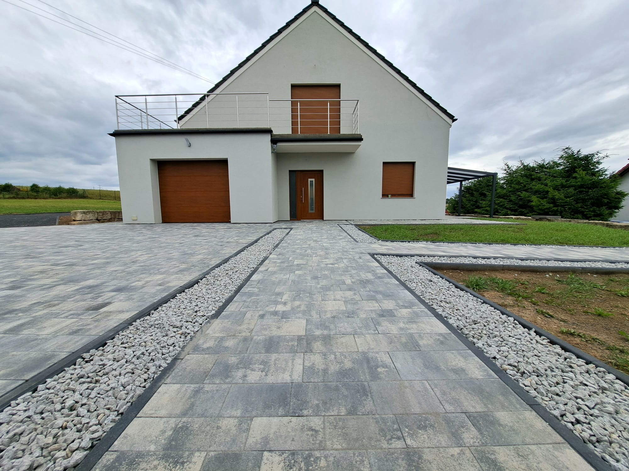 Ułożona szara kostka brukowa z obramowaniem z białych kamieni prowadząca do nowoczesnego domu z brązowymi drzwiami i bramą garażową, widoczna pergola w tle.