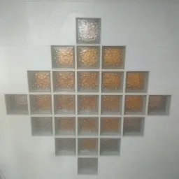 Dekoracyjna ściana z luksferów ułożonych w kształt rombu, z centralnymi kostkami w odcieniach bursztynu i obrzeżami w kolorze srebrzystym, na tle jasnej, prawdopodobnie łazienkowej, ściany.