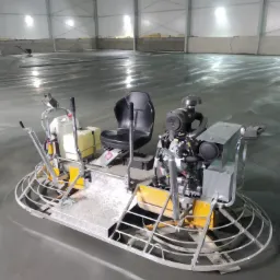 Zacieraczka mechaniczna siedząca na świeżo wylanej, mokrej posadzce w hali przemysłowej z widocznymi śladami po poprzednim przejeździe urządzenia.