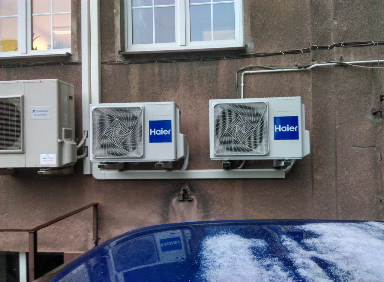 Zewnętrzne jednostki klimatyzacyjne Haier i Fuji Electric zamontowane na elewacji budynku, widok z dołu z fragmentem zaśnieżonego dachu samochodu w kadrze.