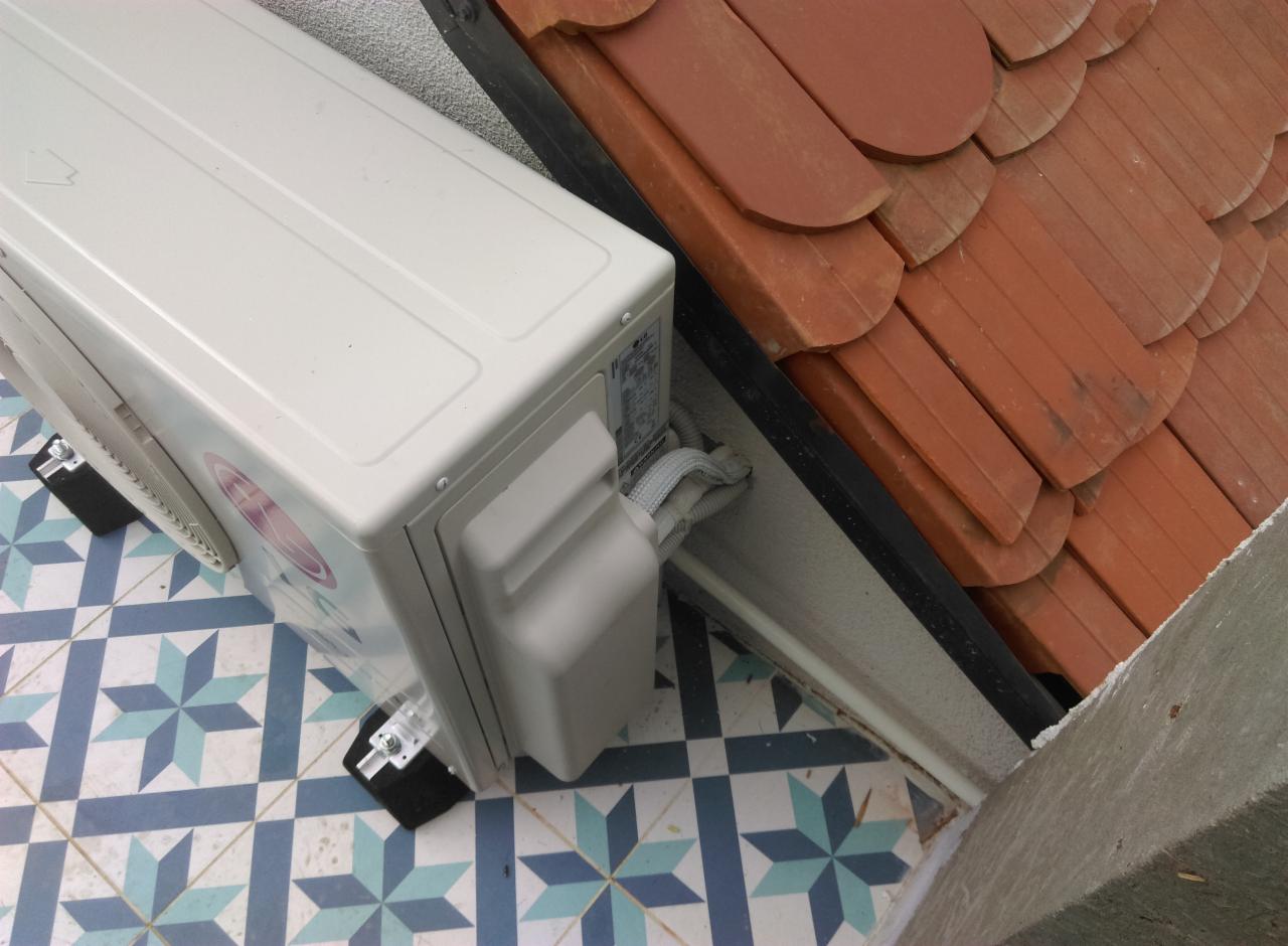 Zewnętrzna jednostka klimatyzatora LG zamontowana na balkonie z wzorzystą, niebiesko-białą podłogą, widoczne kable i rura odprowadzająca skropliny oraz fragment dachu z ceramiczną dachówką.