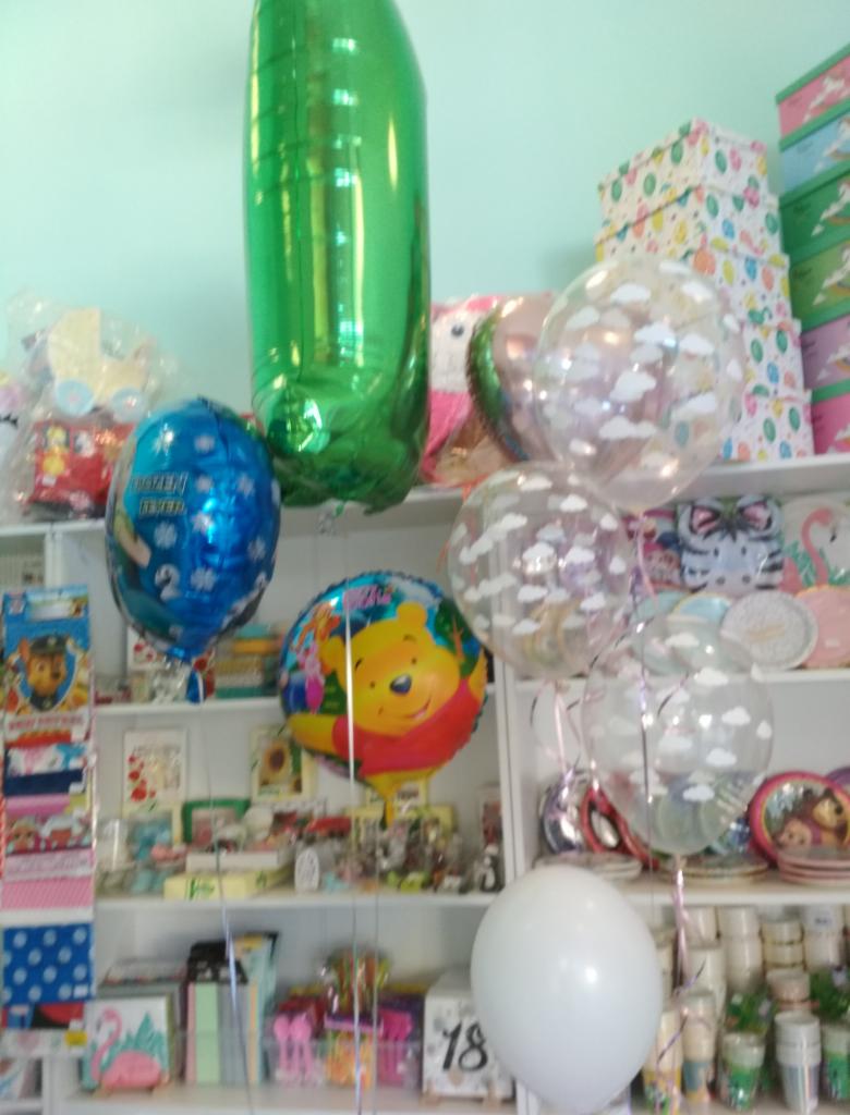 Różnokolorowe balony (zielony, niebieski z motywem zimowym, z wizerunkiem Kubusia Puchatka, przezroczyste z chmurkami, biały) na tle półek z prezentami i dekoracjami.