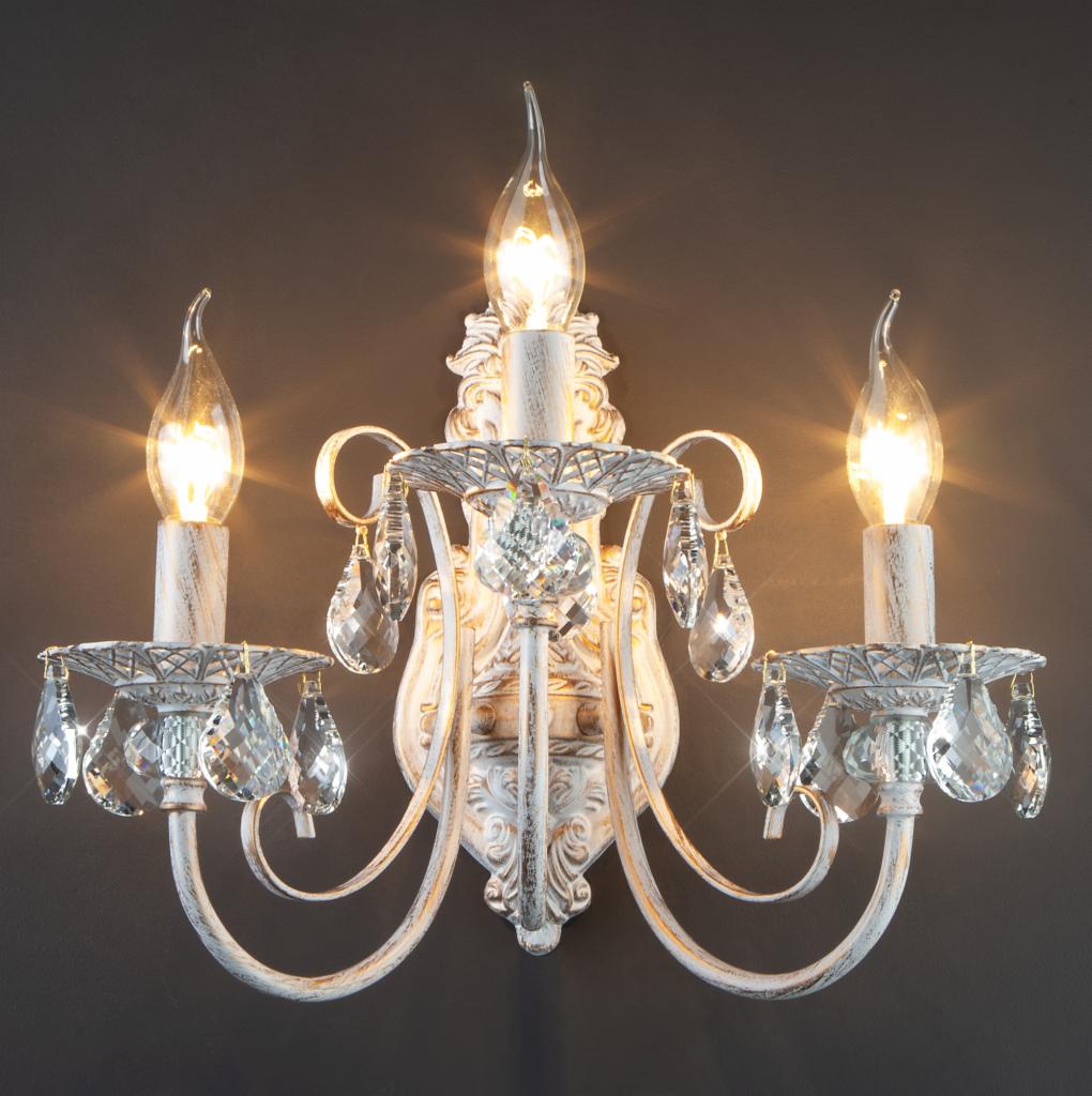 Rustykalny, biały kinkiet z trzema płomieniami świec i kryształowymi zawieszkami na tle szarej ściany.