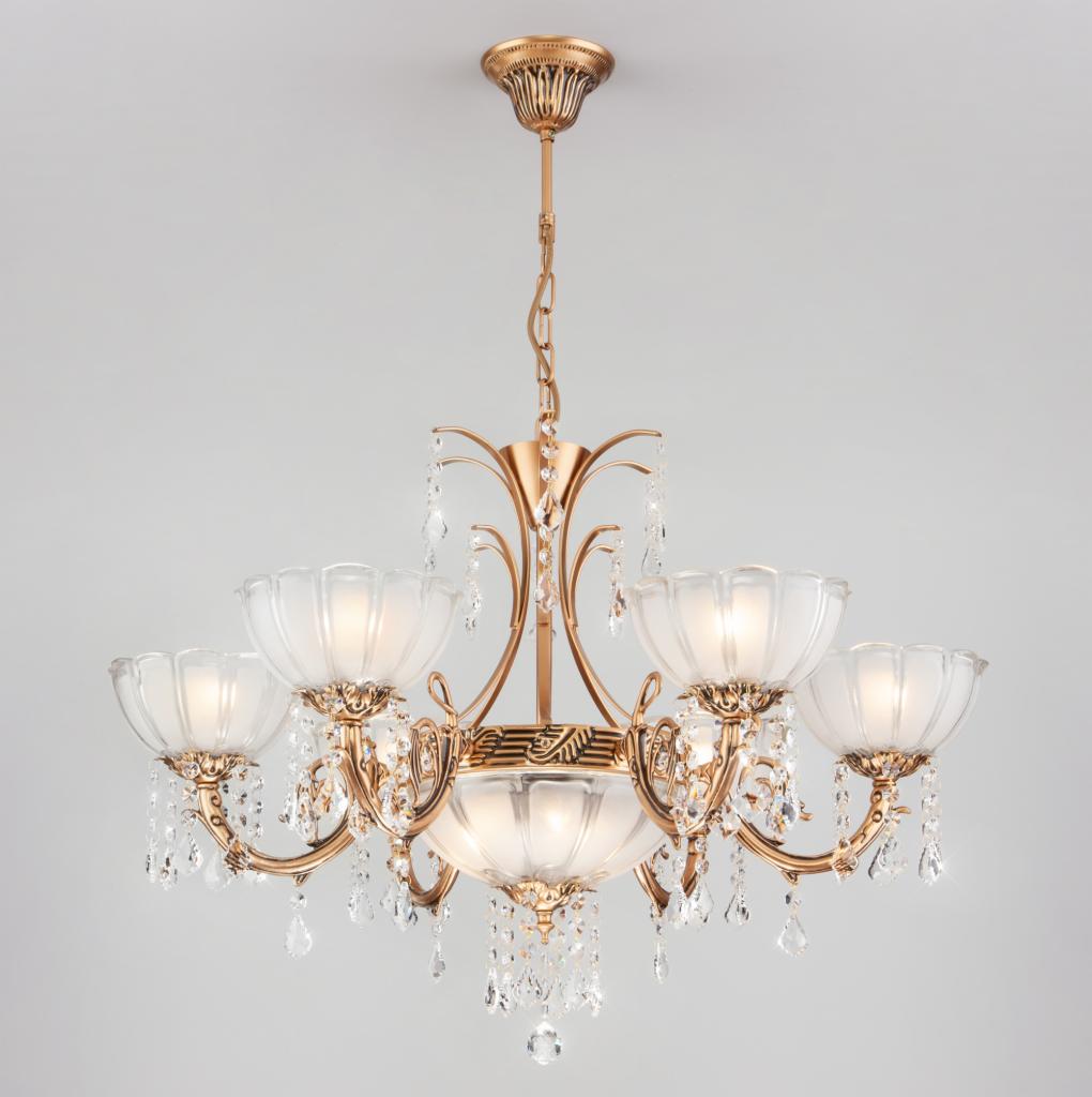 Złoty, sześcioramienny żyrandol z kryształowymi zawieszkami i matowymi, kwiatowymi kloszami, zawieszony na łańcuchu.