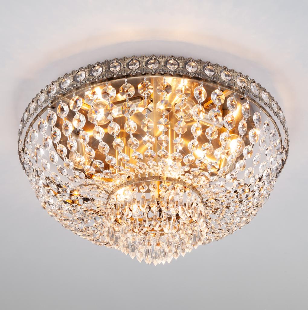 Sufitowa lampa kryształowa z metalową ramą w kolorze starego złota, oświetlająca pomieszczenie ciepłym światłem.