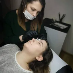 Stylistka w masce i rękawiczkach precyzyjnie aplikuje barwnik na brwi klientki leżącej na białym leżance, widoczne narzędzia na stoliku w tle.