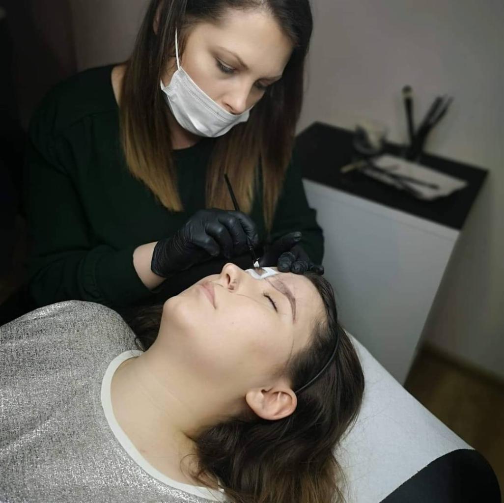 Stylistka w masce i rękawiczkach precyzyjnie aplikuje barwnik na brwi klientki leżącej na białym leżance, widoczne narzędzia na stoliku w tle.