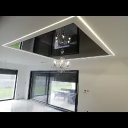 Nowoczesne wnętrze z podwieszanym sufitem o trójkątnym kształcie, podświetlonym listwami LED, oraz kryształowym żyrandolem, widoczne duże okna przesuwne z roletami.