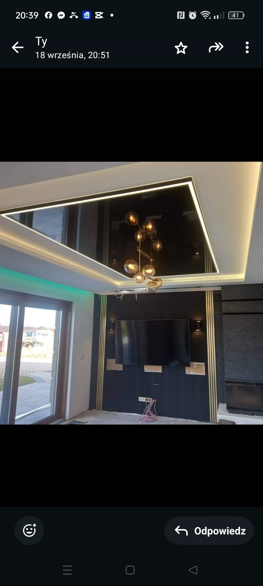 Nowoczesny salon z podwieszanym sufitem z lustrzanym panelem, oświetleniem LED i designerską lampą wiszącą, ściana z ciemnego drewna z złotymi akcentami i miejscem na telewizor, widoczne...