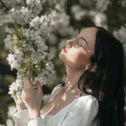 Portret kobiety w okularach, spoglądającej w górę, trzymającej gałązkę kwitnącego drzewa, z promieniami słońca na twarzy.