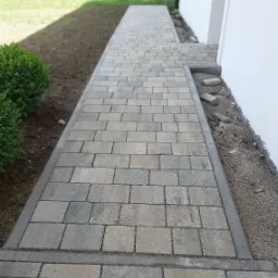 Ułożony chodnik z kostki brukowej w odcieniach szarości, obrzeża z betonu, świeżo obsypane ziemią, przy białej ścianie budynku.