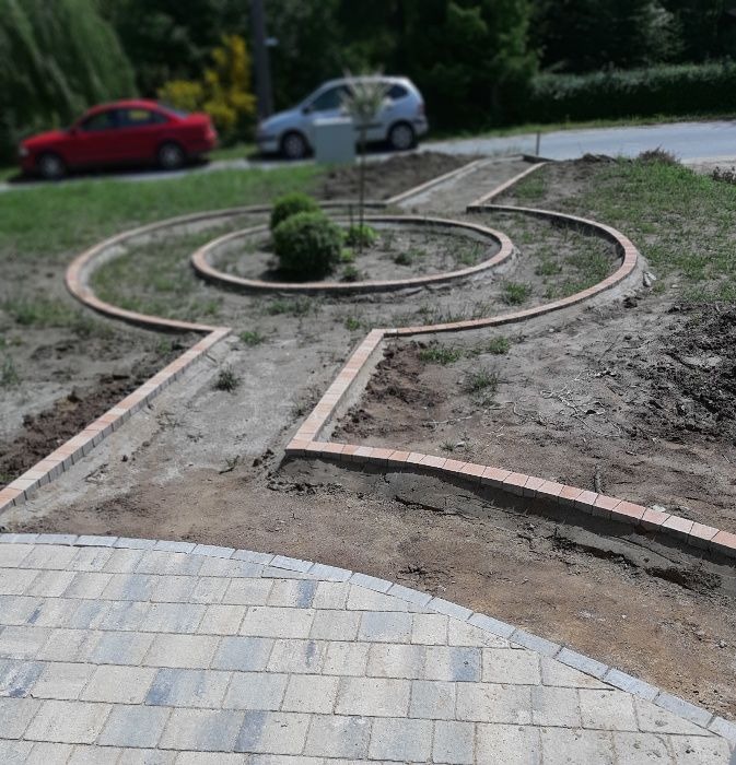 Układanie obrzeży z czerwonej kostki wokół rabaty i ścieżki w ogrodzie, widoczny fragment ułożonej kostki brukowej, w tle samochody i zieleń.