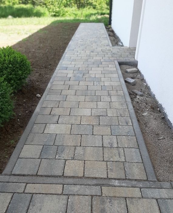 Ułożony chodnik z kostki brukowej w odcieniach szarości, obrzeża z betonu, świeżo obsypane ziemią, przy białej ścianie budynku.