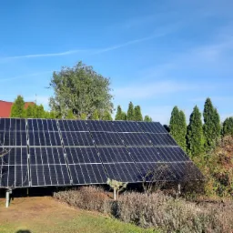 Panel słoneczny na metalowej konstrukcji, otoczony zielenią, w tle rząd wysokich tuj i fragment czerwonego dachu domu pod błękitnym niebem.