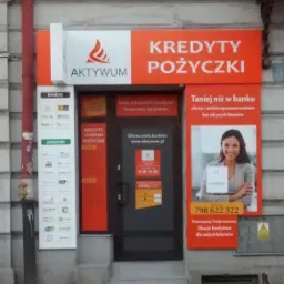Pomarańczowy szyld 'Kredyty Pożyczki' nad wejściem do biura Aktywum, z informacjami o ofercie i godzinach otwarcia, oraz uśmiechniętą kobietą na plakacie.