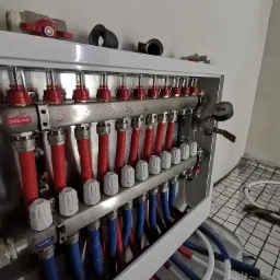 Rozdzielacz instalacji grzewczej podłogowej z rurami doprowadzającymi w kolorach czerwonym i niebieskim, widoczny manometr i napisy 'Zasilanie' i 'Powrót', na podłodze siatka do montażu ogrzewania...