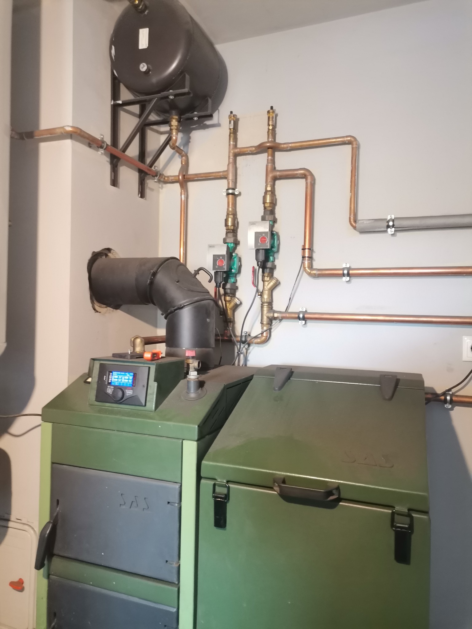 Solidny, zielony piec centralnego ogrzewania marki SAS z podajnikiem, z widocznymi miedzianymi rurami instalacji grzewczej i zasobnikiem ciepłej wody, zamontowany przy szarej ścianie w kotłowni.