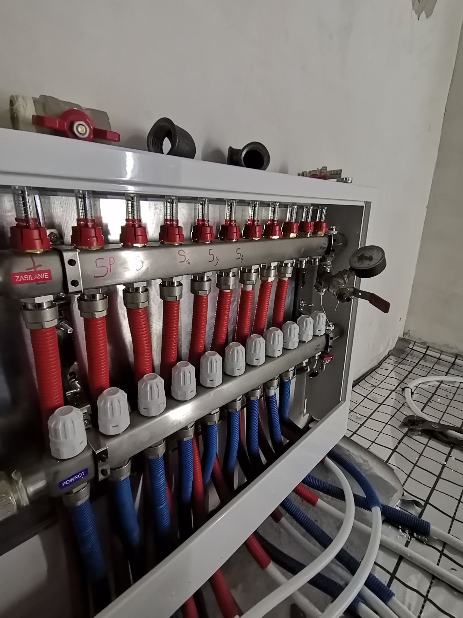 Rozdzielacz instalacji grzewczej podłogowej z rurami doprowadzającymi w kolorach czerwonym i niebieskim, widoczny manometr i napisy 'Zasilanie' i 'Powrót', na podłodze siatka do montażu ogrzewania...