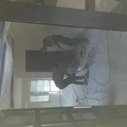 Dwóch mężczyzn w trakcie skuwania starej posadzki w pomieszczeniu zabezpieczonym folią malarską, widok przez framugę drzwi.