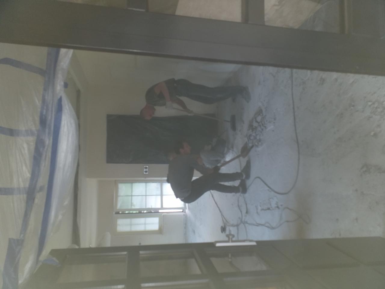 Dwóch mężczyzn w trakcie skuwania starej posadzki w pomieszczeniu zabezpieczonym folią malarską, widok przez framugę drzwi.