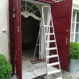 Odrestaurowane, bordowe drzwi wejściowe z ozdobnym nadprożem, widoczne z bliska, z rozstawioną aluminiową drabiną, na zewnątrz budynku z kamienną elewacją.