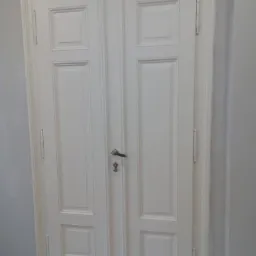 Białe, dwuskrzydłowe drzwi wewnętrzne z dekoracyjnymi panelami i mosiężnym zamkiem, widoczne na tle szarej ściany.