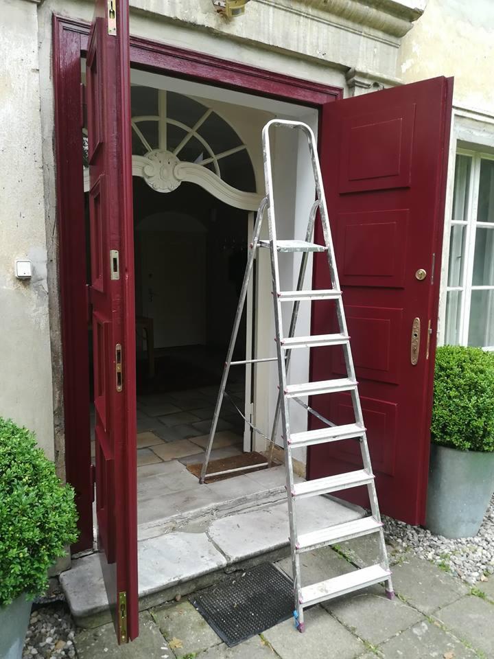 Odrestaurowane, bordowe drzwi wejściowe z ozdobnym nadprożem, widoczne z bliska, z rozstawioną aluminiową drabiną, na zewnątrz budynku z kamienną elewacją.