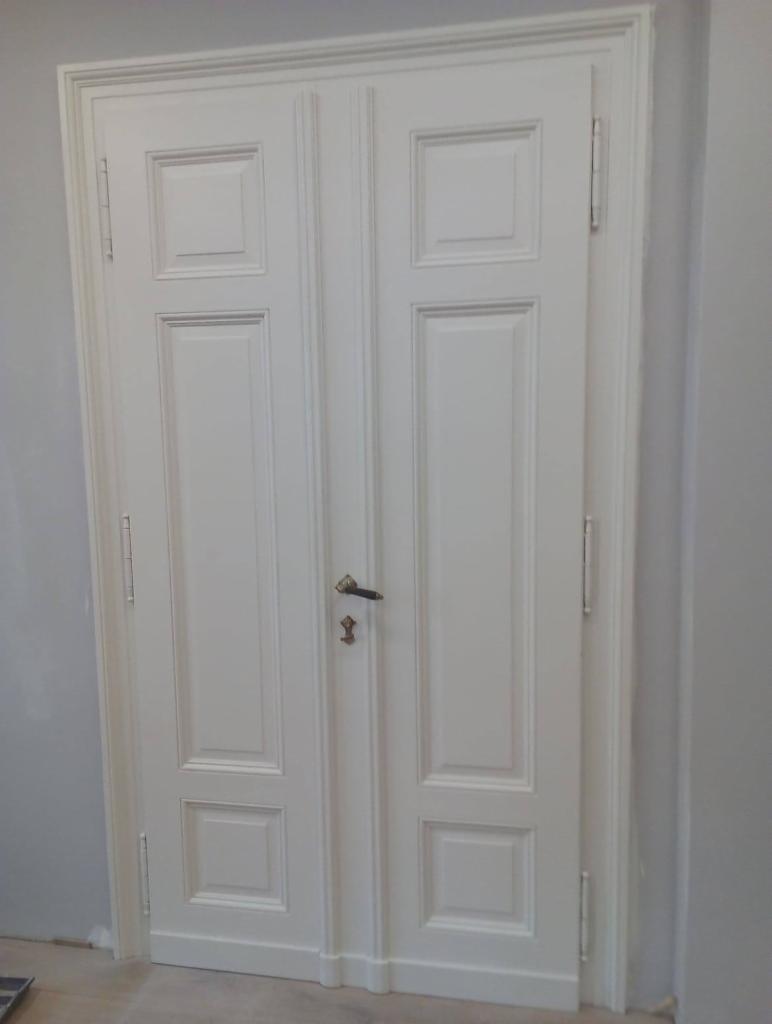 Białe, dwuskrzydłowe drzwi wewnętrzne z dekoracyjnymi panelami i mosiężnym zamkiem, widoczne na tle szarej ściany.