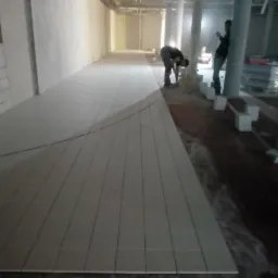 Układanie jasnych płytek podłogowych w długim, słabo oświetlonym korytarzu; widoczni dwaj pracownicy podczas montażu, z zapasem płytek w tle.