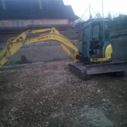 Żółto-zielona mini koparka na grysie, z widoczną ścianą oporową z betonowych elementów ażurowych w tle.