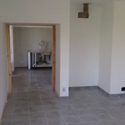 Szare płytki podłogowe w pustym, jasnym pomieszczeniu, widok na częściowo wyposażoną łazienkę z umywalką przez drewniane futryny drzwi.