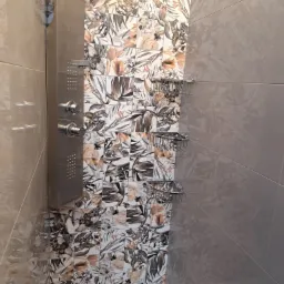 Wykończona kabina prysznicowa z panelami z motywem florystycznym i półkami drucianymi, widoczne baterie i fragment podłogi z folią ochronną.