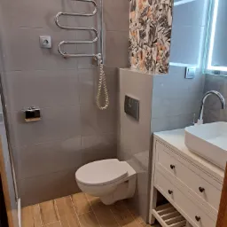 Nowoczesna łazienka z szarymi płytkami, drewnopodobną podłogą, wiszącą toaletą, grzejnikiem drabinkowym, umywalką nablatową na białej szafce z szufladami oraz lustrem z podświetleniem LED...