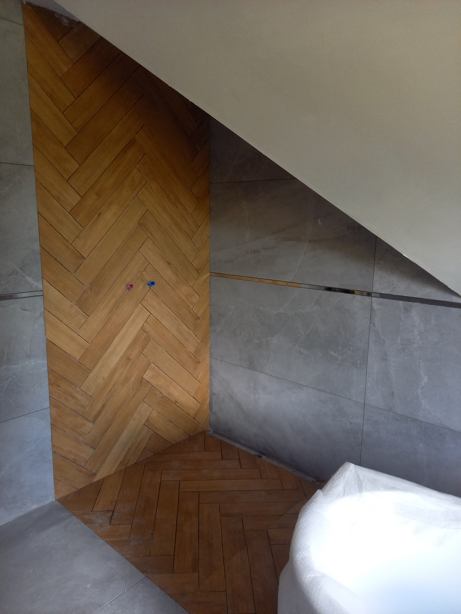 Ukośne ułożenie parkietu w jodełkę na ścianie i podłodze łazienki, z szarymi płytkami imitującymi kamień i fragmentem wanny zabezpieczonej folią.