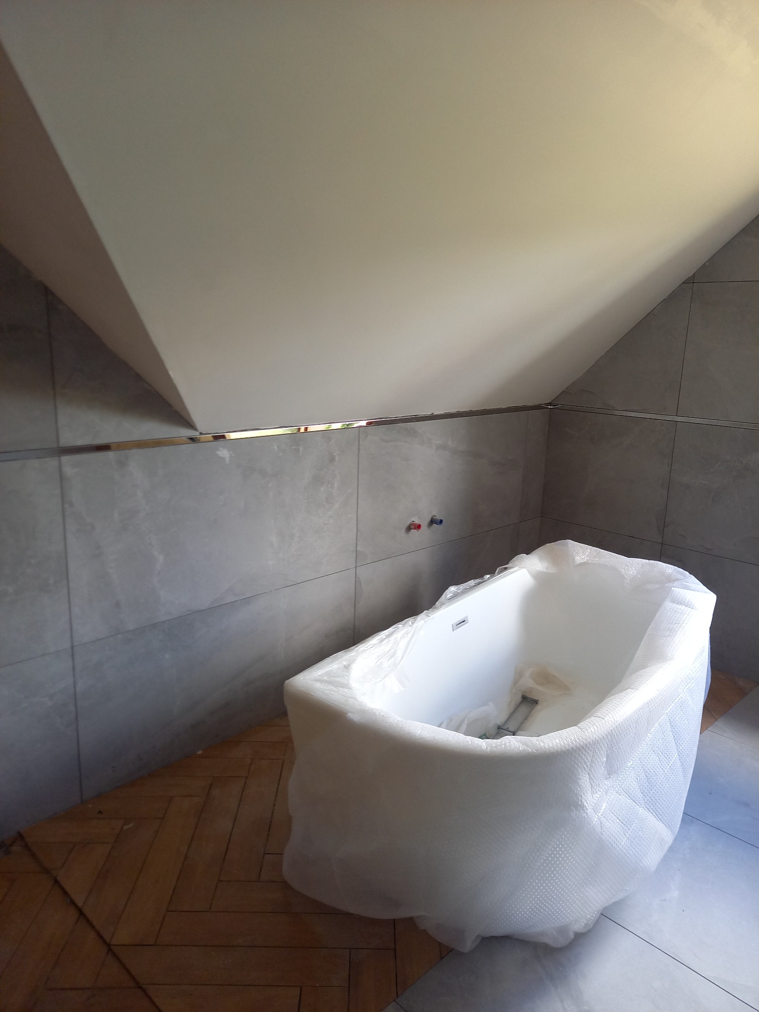 Nowa, biała wanna owinięta folią ochronną stoi w łazience z szarymi płytkami na ścianach i drewnianą podłogą w jodełkę, widoczne przyłącza wodne na ścianie.