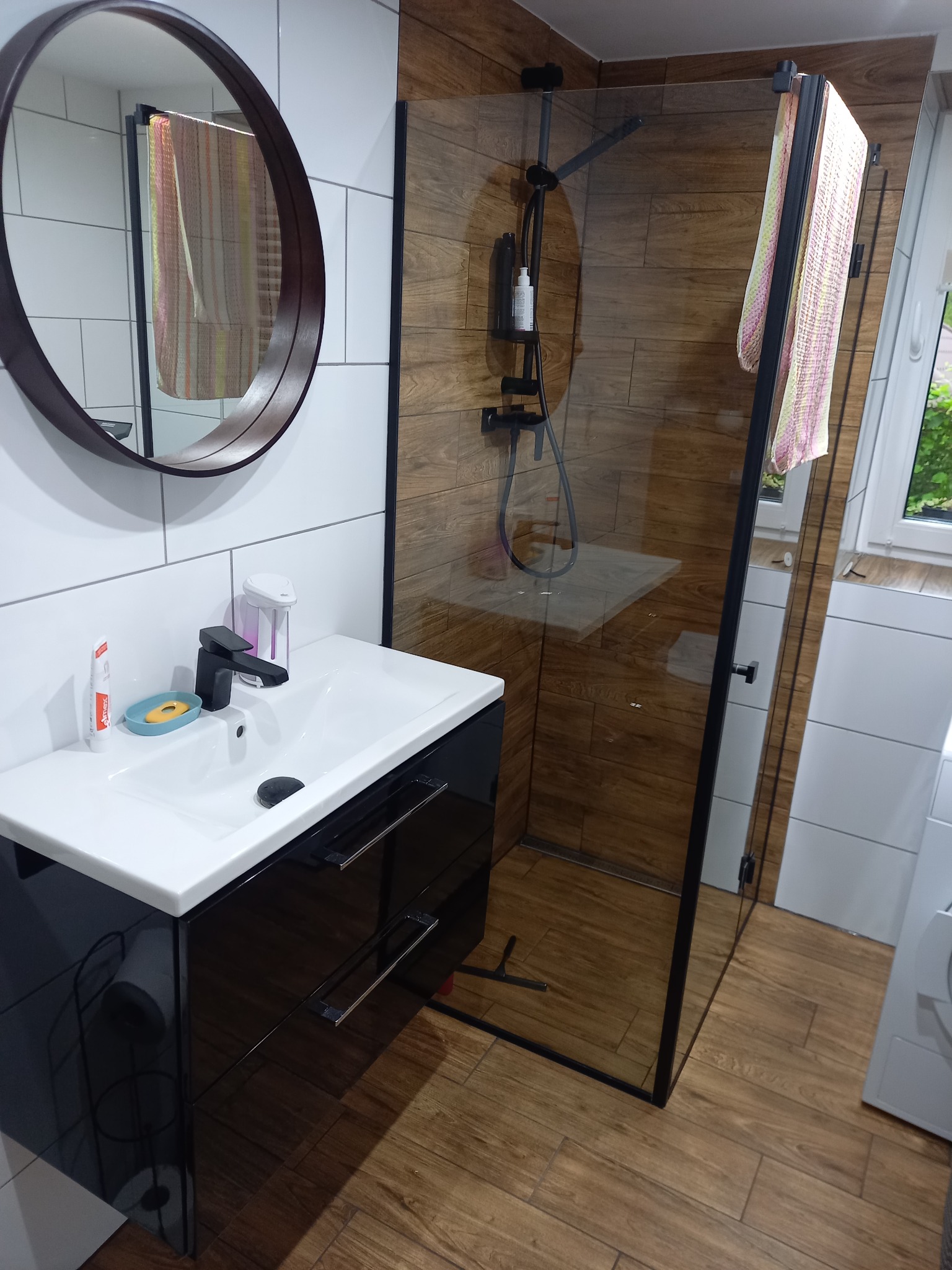 Nowoczesna łazienka z kabiną prysznicową typu walk-in, umywalką nablatową na szafce z czarnym połyskiem, okrągłym lustrem i drewnopodobnymi płytkami.