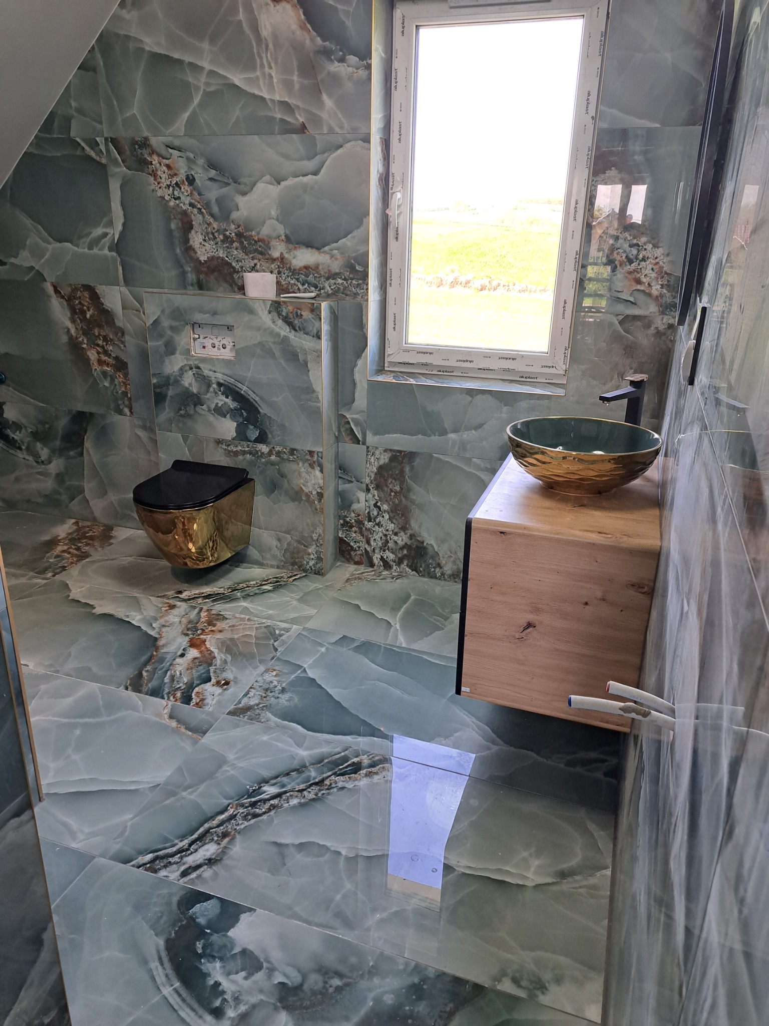 Wykończona łazienka z płytkami imitującymi marmur w odcieniach szarości i zieleni, wisząca toaleta w kolorze złotym, umywalka w kształcie misy z drewnianą szafką pod oknem.