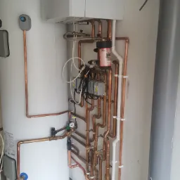 Biały bojler i piec gazowy Vaillant z licznymi miedzianymi rurami, zaworami i elementami instalacji, z widocznym filtrem magnetycznym na ścianie.