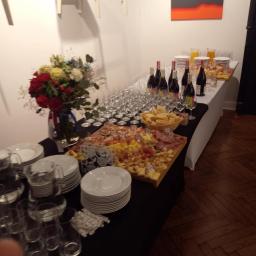 Flomaro sp.zo.o. - Stół cateringowy z deską serów, wędlin i owoców, kieliszki do wina, szklanki z sokiem, butelki wina, talerze, dekoracja kwiatowa, w tle obraz z abstrakcyjnym motywem.