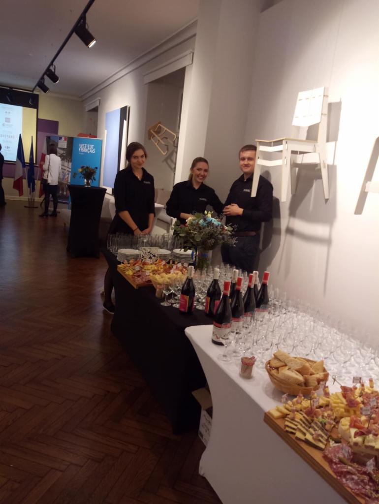 Personel obsługujący stół z przekąskami, winem i szkłem podczas wydarzenia w galerii sztuki z flagami w tle.