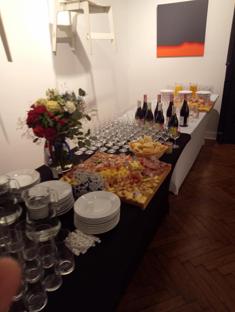 Stół cateringowy z deską serów, wędlin i owoców, kieliszki do wina, szklanki z sokiem, butelki wina, talerze, dekoracja kwiatowa, w tle obraz z abstrakcyjnym motywem.