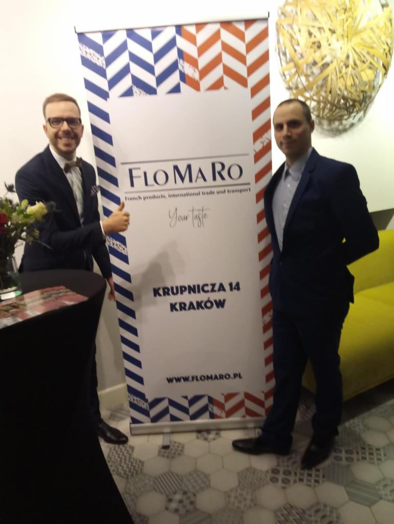 Dwaj mężczyźni w garniturach pozują obok roll-upu z logo firmy Flomaro, oferującej francuskie produkty oraz transport międzynarodowy, na tle wzorzystej podłogi i dekoracji ściennej.